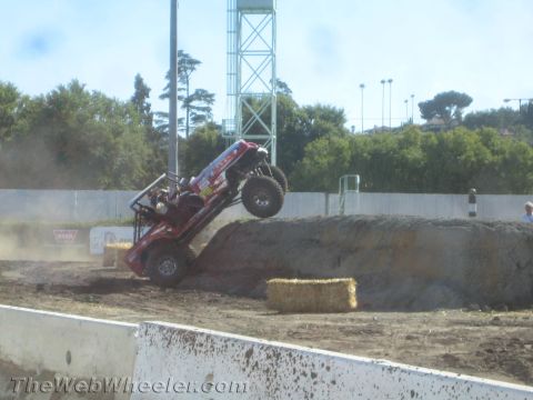 1990 Ford Bronco - Red tube-bodied Jeep off-road buggy climbing steeply nose-up over a dirt mound at an outdoor competition.