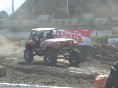 1990 Ford Bronco - Red lifted Jeep CJ rock crawler with two occupants driving across a dusty dirt course past a Skyjacker banner.
