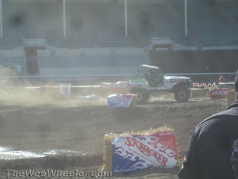 1990 Ford Bronco - Green tube-chassis off-road buggy raising dust on a stadium dirt course past sponsor hay bales, with a spectator silhouette in the foregrou…
