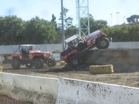 1990 Ford Bronco - Red tube-bodied Jeep off-road buggy climbing a dirt mound while another red Jeep waits at left on the dusty competition course.