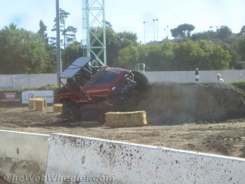 1990 Ford Bronco - Red tube-bodied Jeep off-road buggy tilted nose-down coming off a dirt mound past hay bales at an outdoor competition.