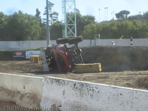 1990 Ford Bronco - red off-road competition truck lies rolled on its side in a dirt arena next to scattered hay bales and a concrete barrier.