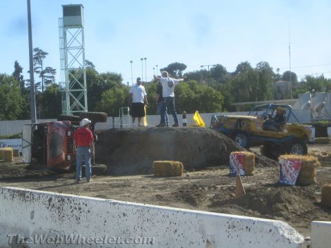 1990 Ford Bronco - People stand atop a dirt obstacle mound between an overturned red truck and a yellow tube-frame off-road buggy in a competition arena.
