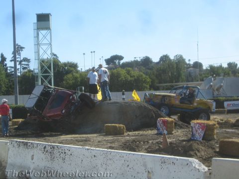 1990 Ford Bronco - red off-road truck climbs a dirt obstacle mound as a yellow tube-frame buggy waits and spotters watch in a fairgrounds arena.