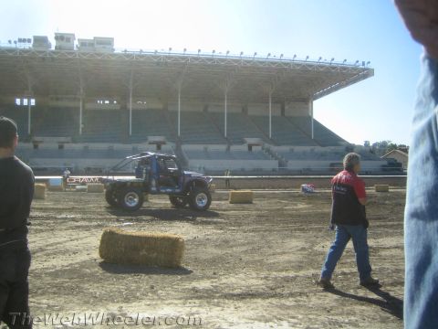 1990 Ford Bronco - lifted off-road truck drives a dirt course in front of a large empty grandstand as people stand by hay bales.