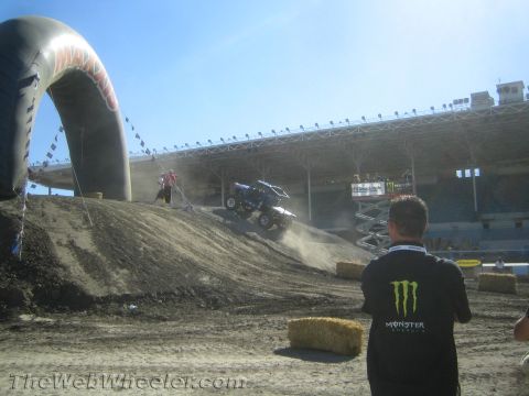 1990 Ford Bronco - off-road buggy crests a dirt jump beneath an inflatable arch as a spectator in a Monster Energy shirt watches in a stadium.