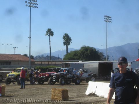 1990 Ford Bronco - lineup of colorful off-road race trucks and buggies sits in a dirt pit area with palm trees and mountains in the background.