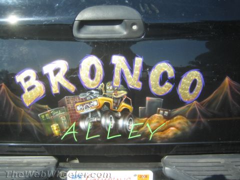 1990 Ford Bronco - dark vehicle tailgate airbrushed with the words BRONCO ALLEY in gold and a cartoon monster-truck creature graphic.