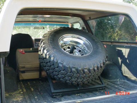1984 Ford Bronco - Spare tire mounted in the cargo area of a light-colored SUV, with a cooler visible behind it.
