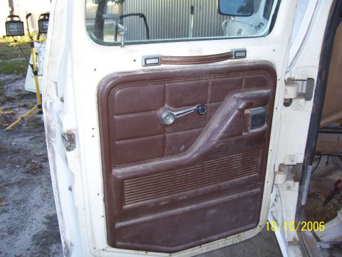 1978 Ford Bronco - Inner face of an open white truck door showing a dusty brown vinyl door panel with manual window crank, molded armrest with pull handle, an…