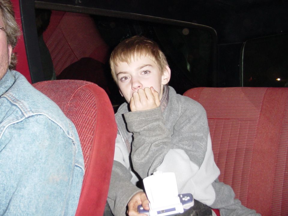 Kid in the back seat of a vehicle, holding a small white object. Another person is visible on the left