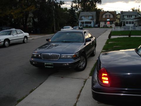 2000 Ford Crown Victoria - A dark blue sedan is parked on the street in front of houses, with another vehicle visible in the foreground.