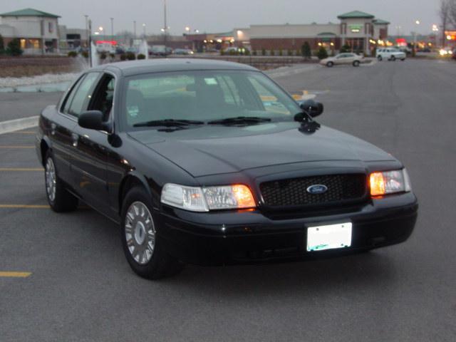 Front three-quarter view of a black Ford Crown Victoria sedan in a parking lot with turn signals lit