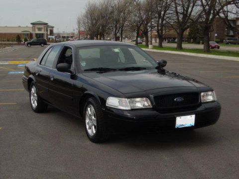 2003 Ford Crown Victoria - This black Ford Crown Victoria is parked in a lot with trees and buildings in the background.