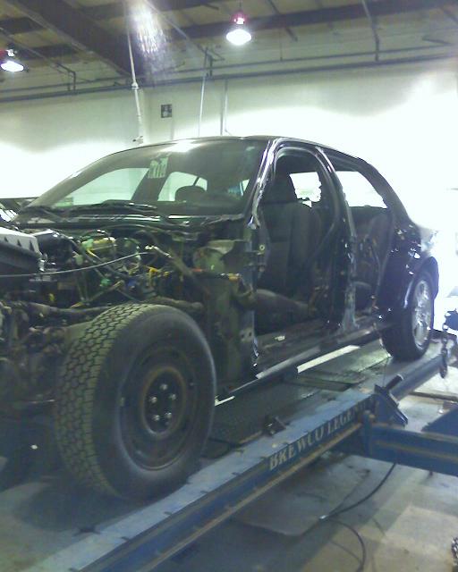 Side view of a heavily damaged car on a lift, stripped of body panels