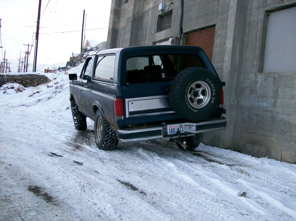 Dark blue SUV parked on a snowy road, rear view. Spare tire on back, license plate reads 180-NIS