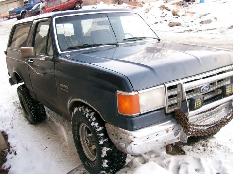 1988 Ford Bronco - Ford Bronco in snow, modified with large tires and a front bumper chain. Other vehicles in background.