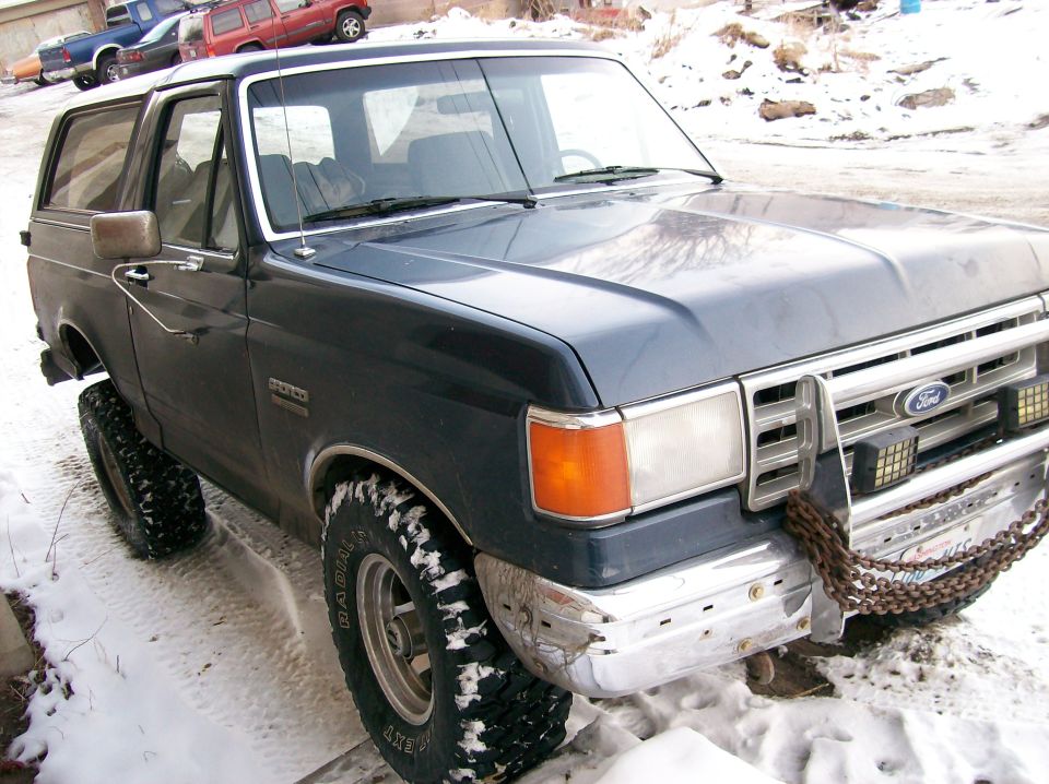 Dark blue Ford Bronco with large tires and a chain on the front bumper, parked in snow. Other vehicles are visible in the background