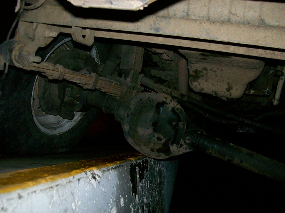 Underside view of a vehicle's rear axle, leaf springs, and tire. The axle housing is round and rusty