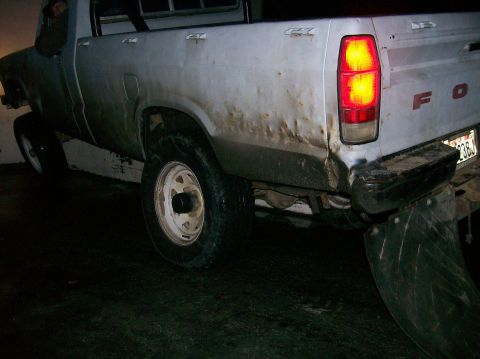1988 Ford Bronco - Rear view of a weathered white pickup truck, showing damage and mud flaps, with its taillight illuminated.