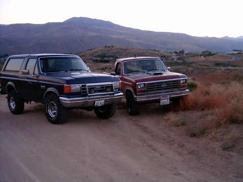 1988 Ford Bronco - Two classic Ford trucks, a Bronco and an F-Series, are parked side-by-side on a dusty road.