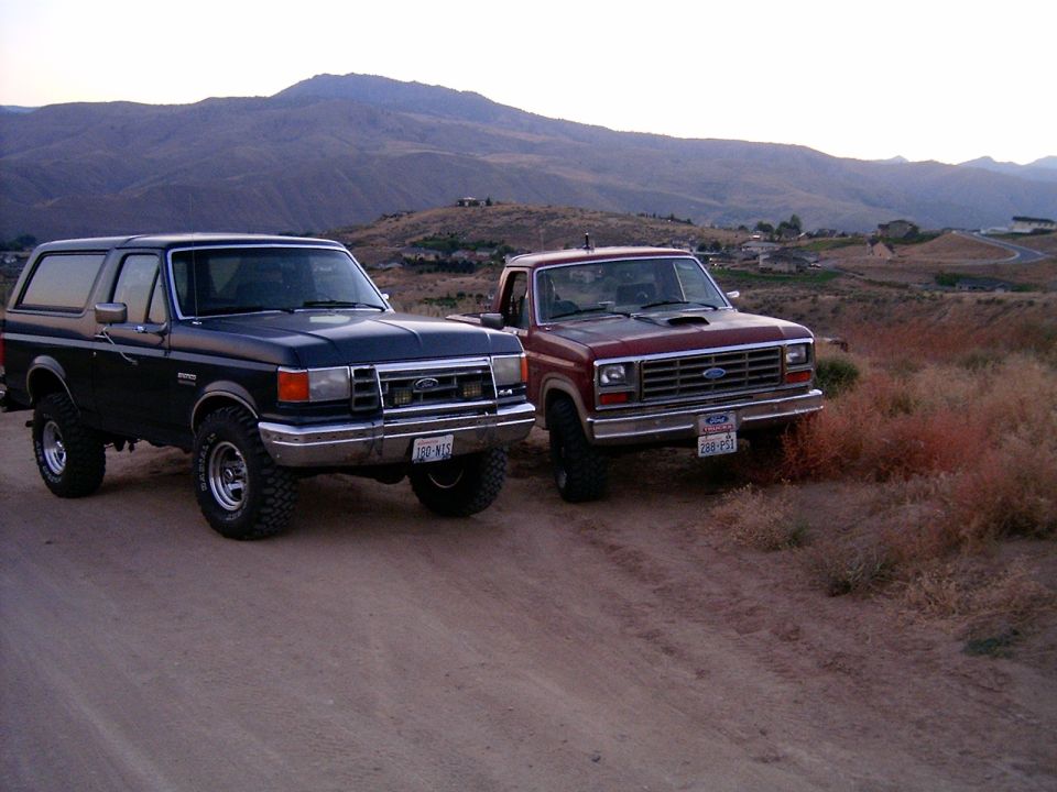 Black Ford Bronco and red Ford F-Series truck parked on a dirt road with hills in the background