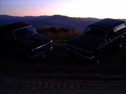 1988 Ford Bronco - Two SUVs are parked on a dirt road as the sun sets behind rolling hills.