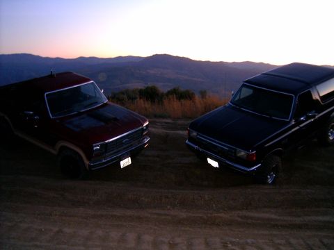 1988 Ford Bronco - Two 1988 Ford Broncos are parked on a dirt hill overlooking a mountain range at sunset.