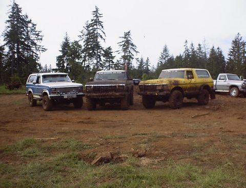 1985 Ford Bronco - Several off-road vehicles are gathered on a dirt lot with a wooded area behind them.