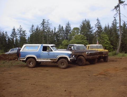 1985 Ford Bronco - Three trucks, including a blue and white Ford Bronco, are parked on a dirt lot with a forest behind them.