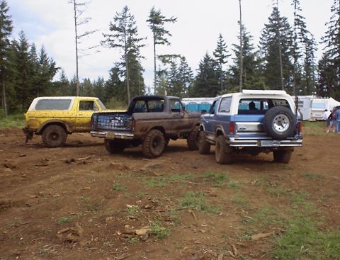 1985 Ford Bronco - Several Ford Broncos are parked on a muddy track, with trees and portable toilets visible in the background.