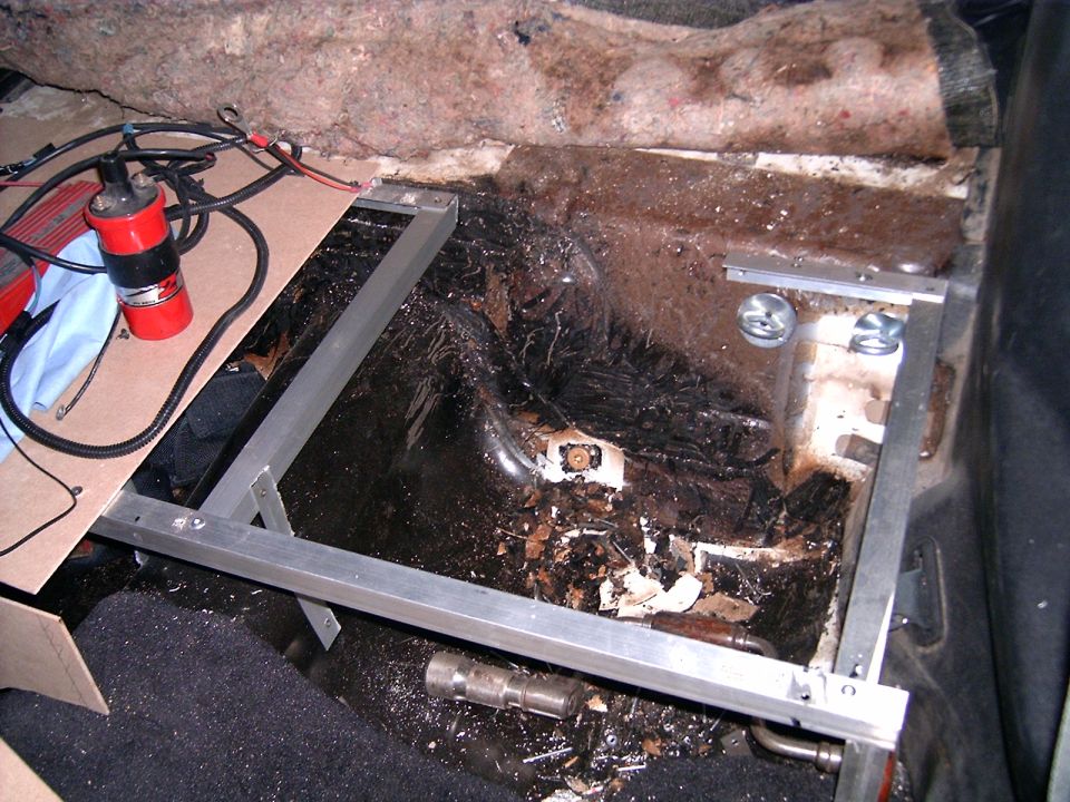 Car interior showing damaged floor, exposed wiring, and metal brackets installed near the rear seat area. A red ignition coil is visible