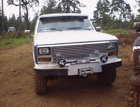 1985 Ford Bronco - A white Ford Bronco is parked on a dirt road, with another vehicle partially visible to its right.