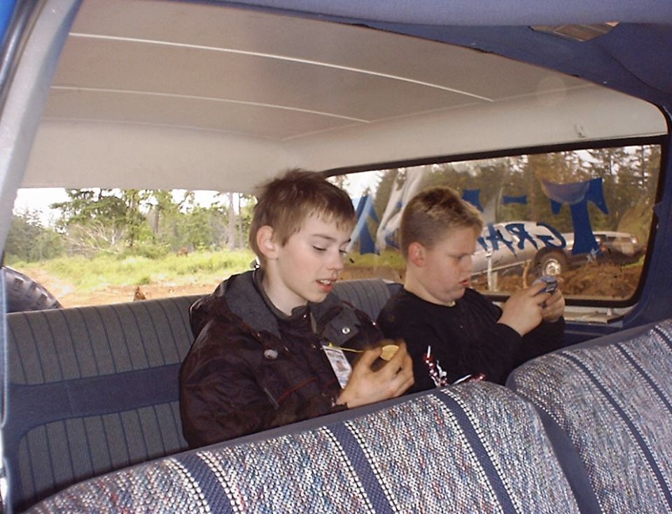 Two boys sit in the back seat of a vehicle, one eating and the other looking at a phone. Trees and another vehicle are visible through the window