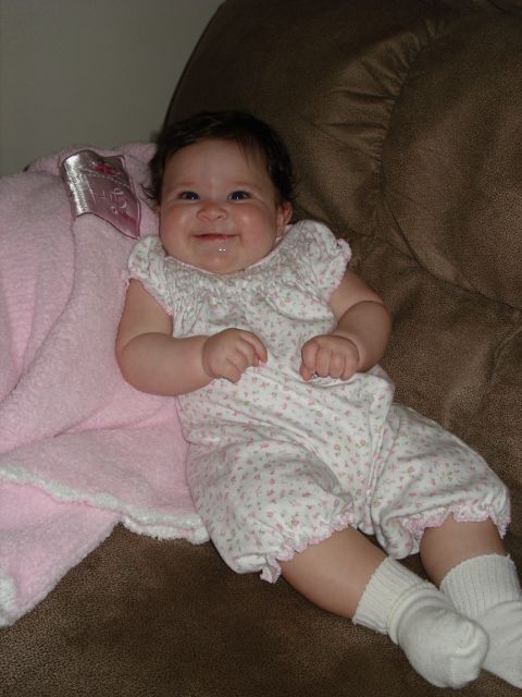 2000 Ford Crown Victoria - A smiling baby girl wearing a floral romper and white socks is sitting on a brown couch, with a pink blanket beside her.