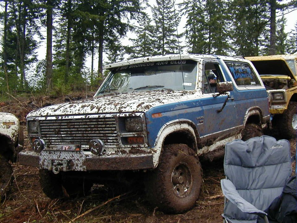Mud-covered blue and white Ford Bronco with large tires, parked in a wooded area next to a yellow vehicle