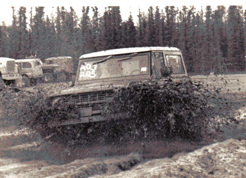 Muddy SUV plowing through mud bog with military trucks and forest behind