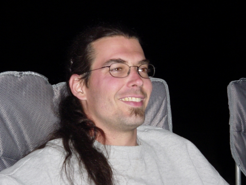Smiling man with long ponytail and glasses seated indoors at night