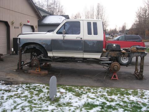 1990 Ford F-250 - This 1990 Ford F-250 is undergoing repairs, with its chassis elevated on jack stands. A red pickup truck is visible in the background.