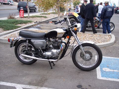 1972 Ford Ranchero - A Triumph Bonneville motorcycle is parked in a lot, with other people and vehicles visible in the background.