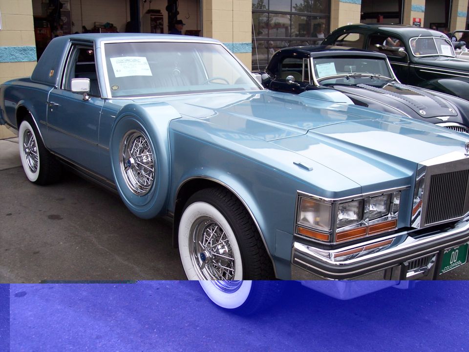 Light blue coupe with wire wheels and a fender-mounted spare tire. Another car is parked behind it