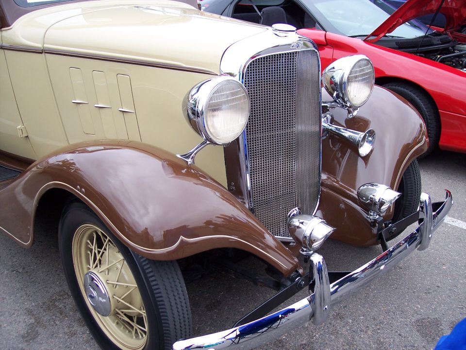 Front end of a two-tone brown and tan vintage car with a chrome grille and round headlights. A red car is parked behind it