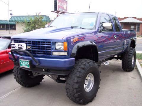 1972 Ford Ranchero - A custom purple pickup truck with large off-road tires and flame graphics is parked next to a red sports car.