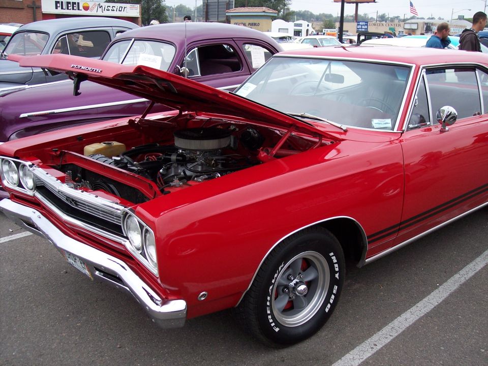 Red muscle car, hood open, showing engine. Another purple car is parked next to it