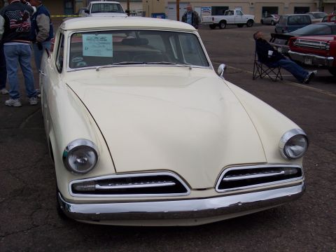 1972 Ford Ranchero - A cream-colored Studebaker pickup truck is parked outside, with people standing nearby and a man sitting in a chair in the background.