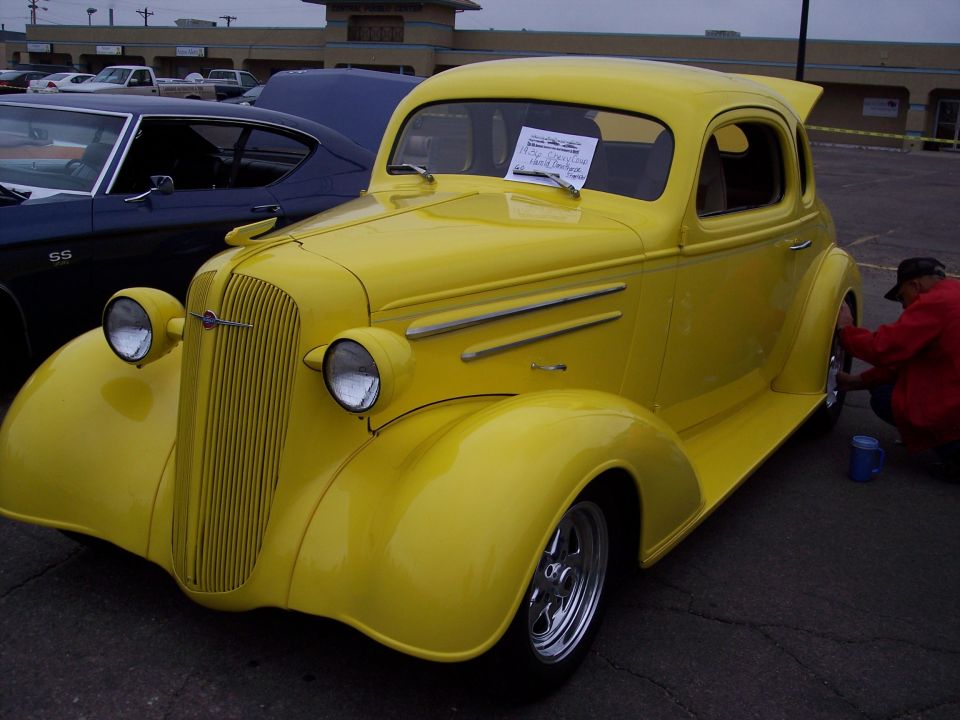 Yellow hot rod coupe with a blue car behind it. A person in a red jacket is cleaning the wheel