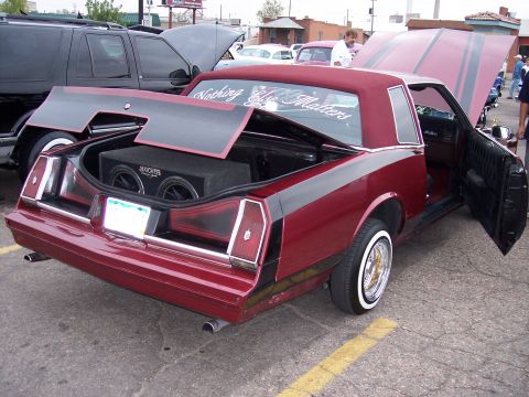 1972 Ford Ranchero - A customized maroon coupe with racing stripes and a lifted trunk displaying subwoofers is parked next to a black SUV.