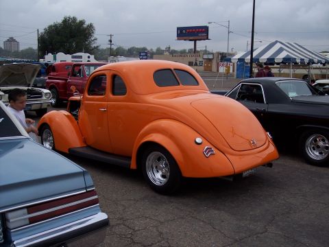 1972 Ford Ranchero - Orange hot rod coupe with rounded rear window, parked next to other vehicles at a show.