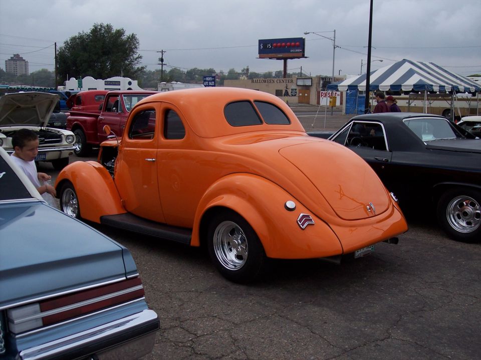 Orange hot rod coupe with rounded rear window, parked next to black El Camino and blue car