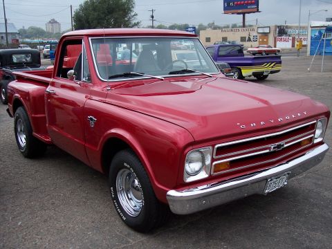1972 Ford Ranchero - A bright red 1967 Chevrolet pickup truck is parked in a lot with other vehicles visible in the background.